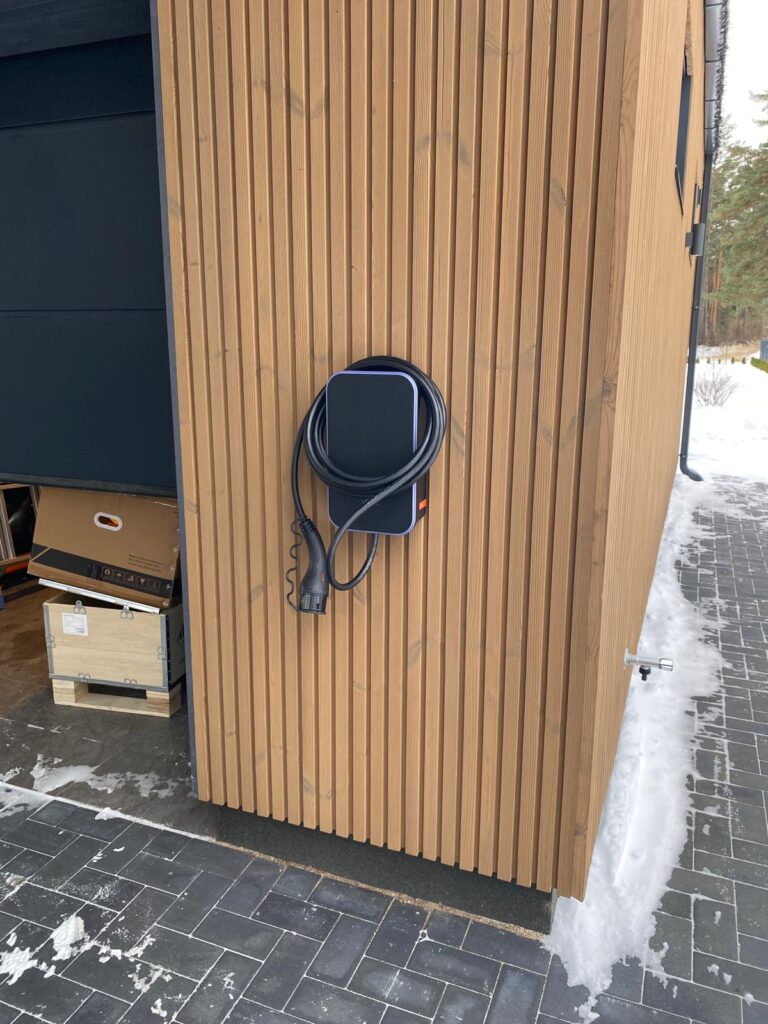 Elektriskās automašīnas lādētājs pie mājas sienas ar koka apdari. Melns lādētājs ar aptītu kabeli.