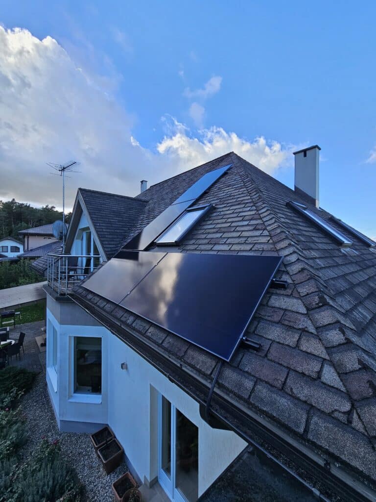 Māja ar saules paneļiem uz jumta un jumta logiem, zilas debesis fonā.