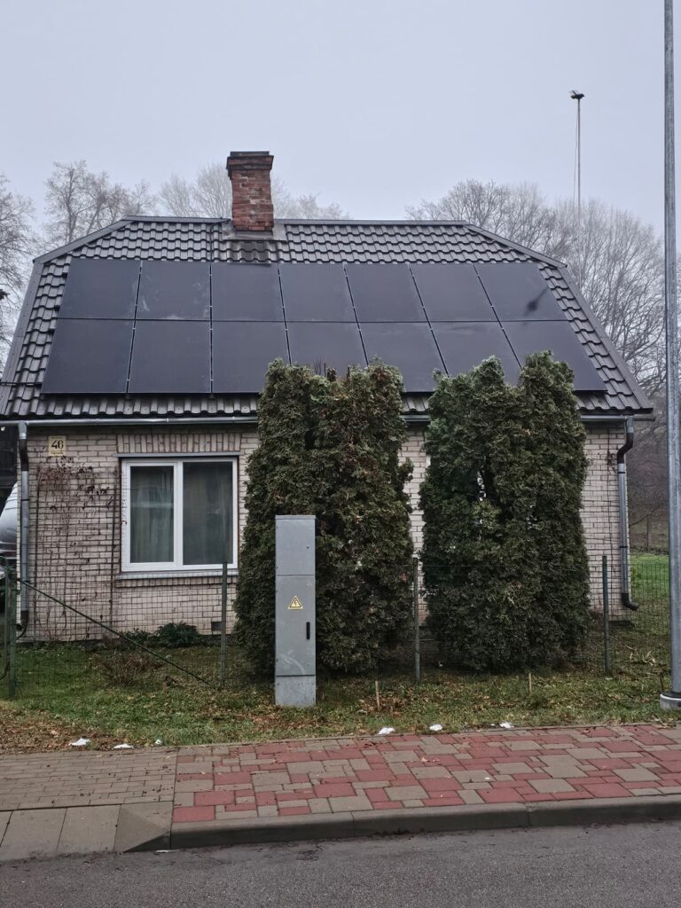 Māja ar saules paneļiem uz jumta, zaļi krūmi priekšplānā un elektrības skapis. Ķieģeļu siena redzama.