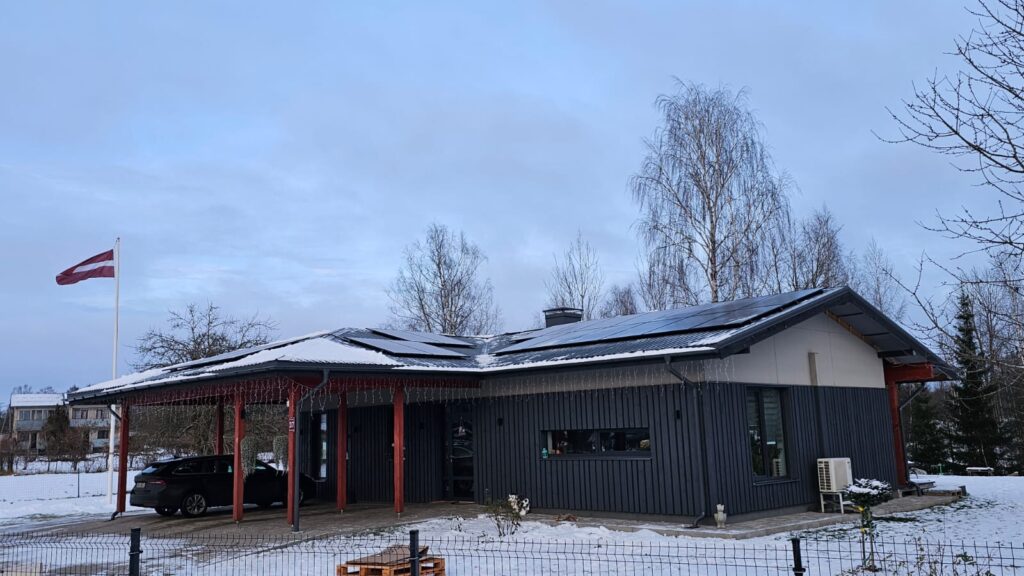 Māja ar saules paneļiem, Latvijas karogs, ziemas ainava. Modernā arhitektūra, tumša fasāde, sniegs.