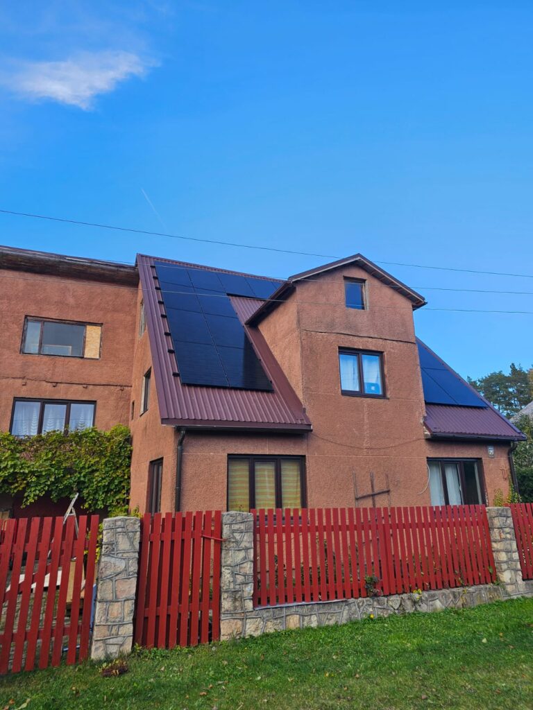 Māja ar saules paneļiem uz jumta, sarkana sēta, zaļš zālājs. Zils debesis fonā.