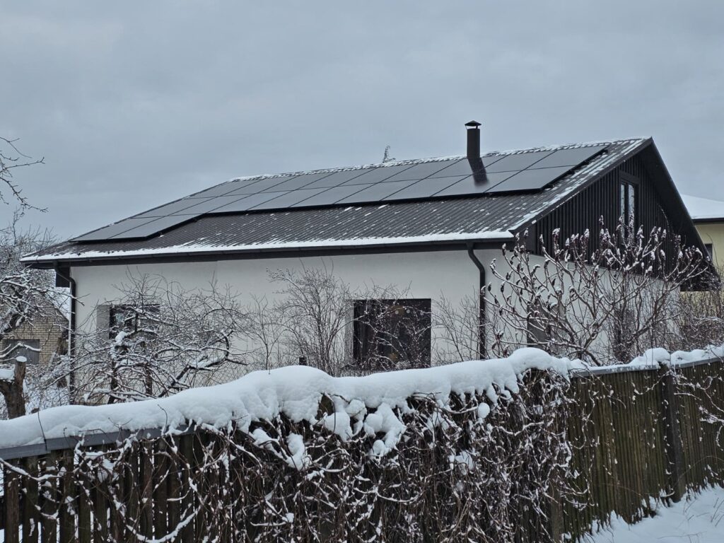 Māja ar saules paneļiem ziemā, sniegs uz žoga. Tumši saules paneļi uz jumta, balta māja.