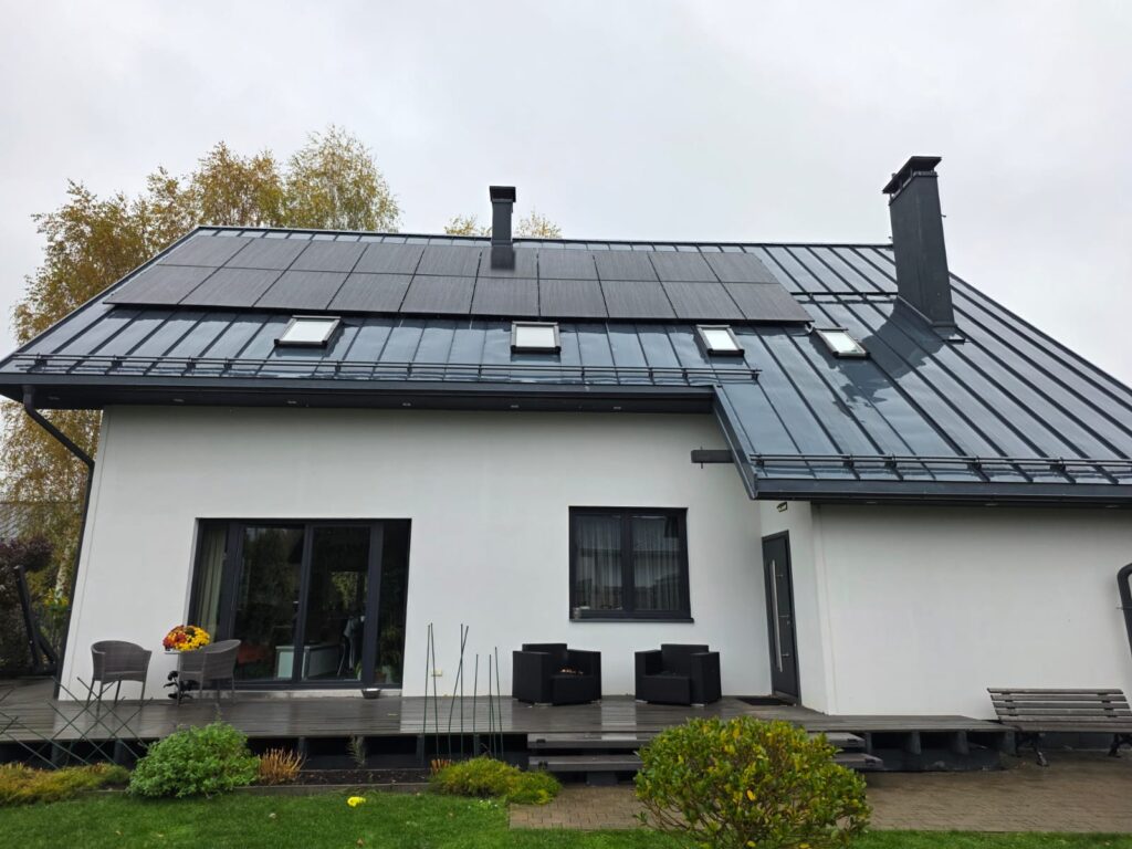 Mūsdienīga māja ar saules paneļiem uz jumta, terasi un dārzu. Balts fasāde, melns jumts.