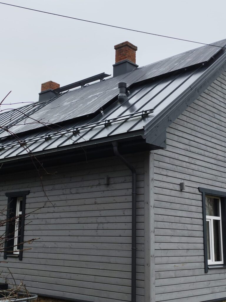 Pelēka koka māja ar metāla jumtu, skursteņiem un saules paneļiem. Lietus notekcaurule pie sienas.