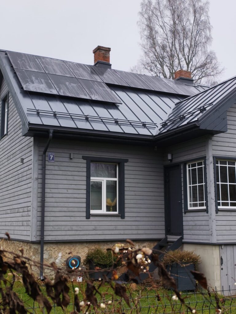 Pelēka koka māja ar saules paneļiem uz jumta. Mājas numurs 7. Ainava pelēkā dienā.