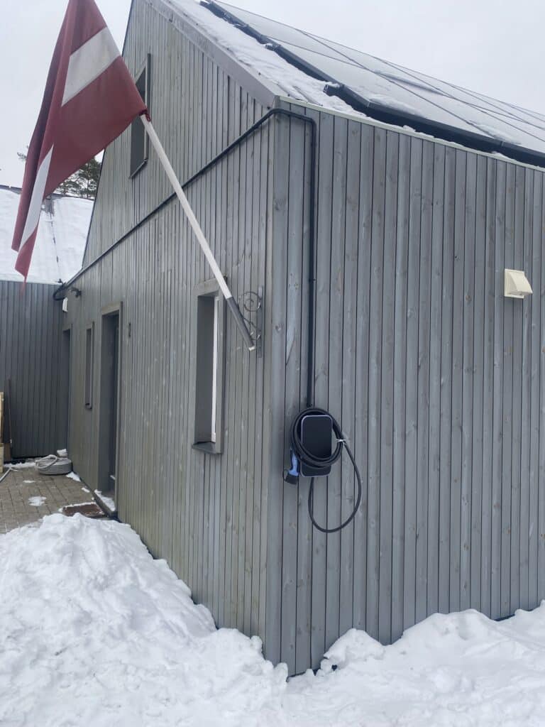 Elektroauto uzlādes stacija pie mājas sienas ar Latvijas karogu. Sniegs uz jumta.