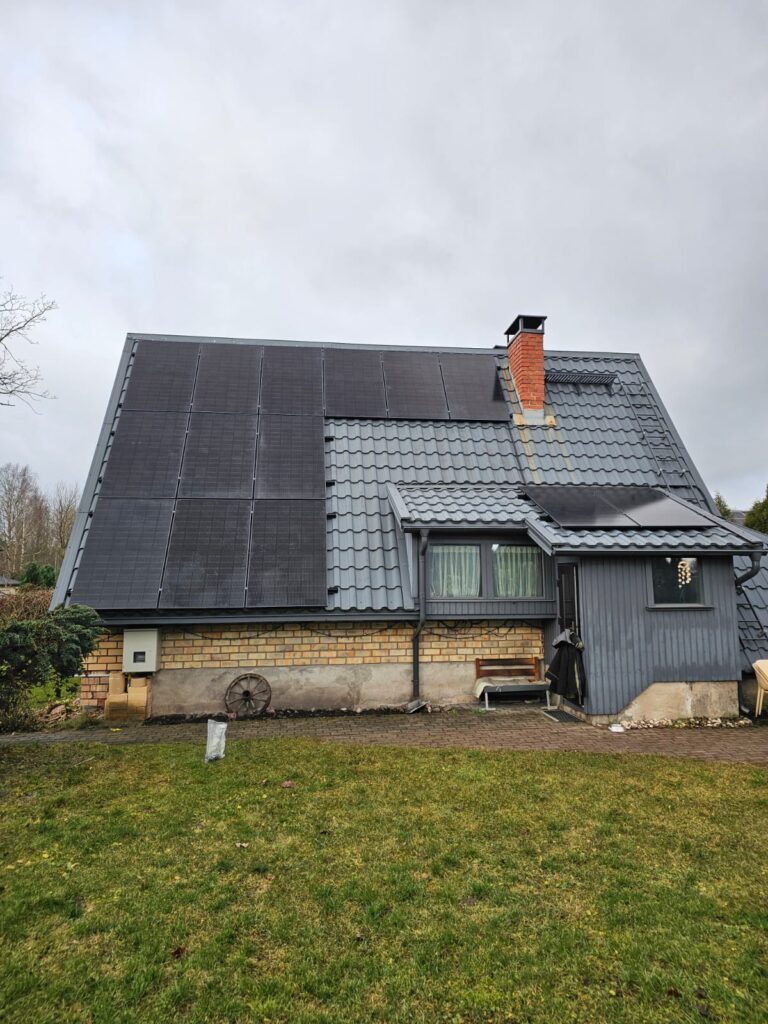 Māja ar saules paneļiem uz jumta, ķieģeļu siena un zaļš zāliens priekšplānā.